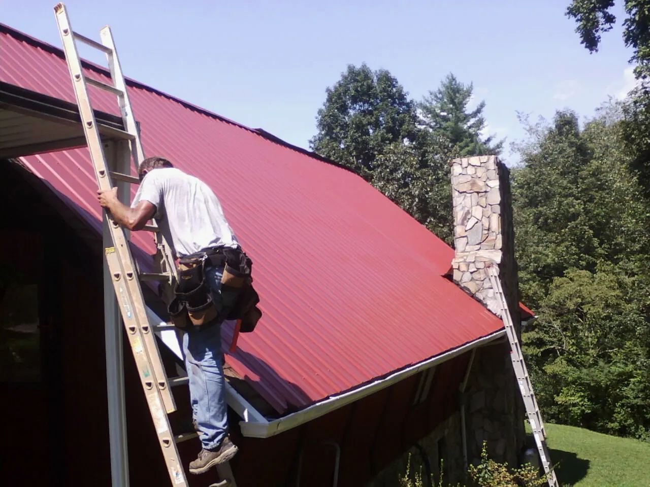 Roofer on a ladder beside a red metal roof and stone chimney — Soffit & Fascia Repair by Roofing Brothers in Canehill